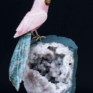 Puede incluir: Una escultura de loro de cuarzo rosa posada sobre una geoda con exterior verde e interior de cristal blanco. El loro tiene una cola de plumas azules y verdes y un ojo marrón. La escultura se exhibe sobre una base de madera roja.