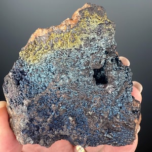May include: A rough, dark gray rock with iridescent blue and gold accents. The rock has a rough, textured surface and a few small holes.