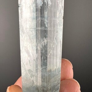 May include: A clear, light blue aquamarine crystal with a rectangular shape. The crystal has a smooth, polished surface and is about 5 cm long.