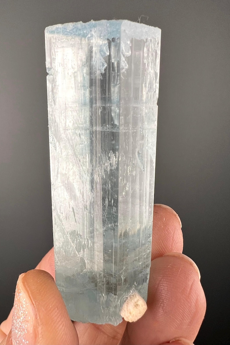 May include: A clear, light blue aquamarine crystal with a rectangular shape. The crystal has a smooth, polished surface and is about 5 cm long.