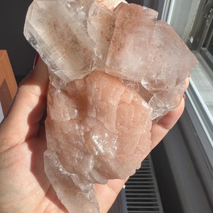 May include: A cluster of translucent, peach-colored crystal formations. The crystals have a rough, textured surface and a natural, organic shape. The light catches the facets, creating highlights and shadows. The overall appearance is of a raw, unpolished mineral specimen.