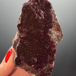 Puede incluir: Un espécimen mineral de color marrón oscuro, con bordes ásperos e irregulares. La superficie está cubierta de pequeños cristales y motas de color más claro. La roca tiene un tono burdeos intenso y un aspecto texturizado.