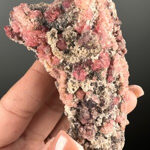 Puede incluir: Un cúmulo de cristales rosados y grises sobre una roca natural rugosa. Los cristales son pequeños y tienen un aspecto brillante y translúcido.