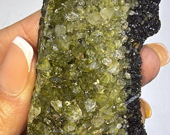 May include: A cluster of green and clear crystals with a black matrix. The crystals are small and have a rough texture. The matrix is smooth and has a dark, almost black color.