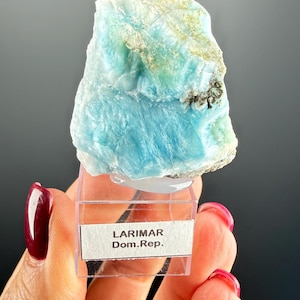 May include: A piece of Larimar gemstone, displaying a mix of blue and white hues. The stone is held in a clear plastic case with a label that reads "LARIMAR Dom.Rep."