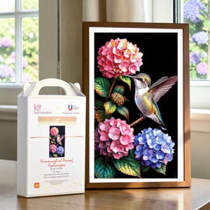 Puede incluir: Un kit de bordado con cuentas con una caja etiquetada Hummingbird Among Hydrangeas junto a un cuadro enmarcado de un colibrí posado en una rama rodeado de hortensias rosas y azules sobre un fondo negro con hojas verdes.