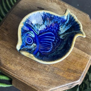 Może przedstawiać: Ceramiczny talerz w kształcie ryby z głębokim niebieskim szkliwem i kremowym obramowaniem. Talerz posiada szczegółowy wzór ryby z teksturowanymi łuskami i płetwami. Talerz jest prezentowany na drewnianej powierzchni.