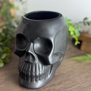 Pode incluir: Um recipiente de cerâmica preto fosco em forma de caveira. O recipiente tem uma abertura larga na parte superior e características faciais detalhadas, incluindo órbitas oculares, uma cavidade nasal e dentes. A caveira é um item decorativo.