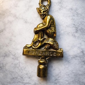 Può includere: Chiave in ottone con una figura di folletto seduto con corna e un volto sorridente. La parola "PENZANCE" è incisa sulla base. La chiave ha un anello in alto e un manico cilindrico.