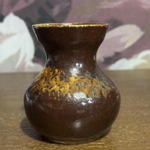 Può includere: Un piccolo vaso in ceramica marrone con corpo arrotondato e collo stretto. Il vaso ha una finitura lucida con un motivo screziato marrone dorato nella metà inferiore. Il bordo è di colore marrone chiaro.