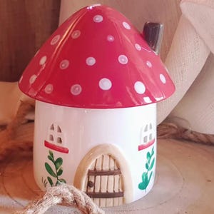 Op de afbeelding: Een grillig keramisch paddenstoelhuisje met een rode kap met witte stippen. Het huis heeft een witte basis, een kleine schoorsteen en een houten deur. Groene bladeren en kleine ramen dragen bij aan de sprookjesachtige charme. Ongeveer 15 cm hoog.