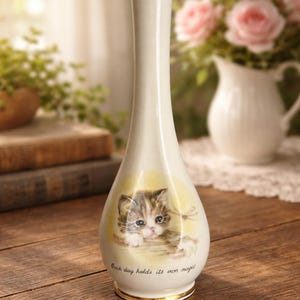 May include: A white ceramic vase with a slender neck and a rounded base. The vase features a painted illustration of a kitten with the text "Each day holds its own magic". A gold band accents the base.