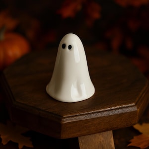 Może przedstawiać: Mała, biała ceramiczna figurka ducha z dwoma czarnymi oczami w kropki. Duch stoi na ciemnobrązowej drewnianej podstawce, z rozmytym tłem z jesiennych liści i dyni. Dekoracja na Halloween.