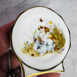 May include: A small, white, scalloped dish with a gold rim. The dish features a painted scene of two rabbits in a field with flowers and a butterfly. The rabbits are brown and white, and the flowers are blue and yellow.