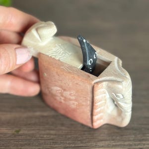 May include: A carved stone box with a lid, featuring a detailed elephant design. The box is primarily beige and reddish-brown, with a black snake-like figure emerging from within. The box is held by a hand.