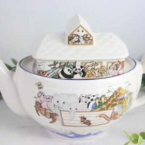 Könnte beinhalten: Weiße Teekanne aus Keramik mit einem hausförmigen Deckel. Die Teekanne ist mit einer Szene von Tieren auf Noahs Arche verziert, darunter ein Panda, eine Giraffe, ein Elefant und ein Schwein. Der Deckel hat ein kleines Fenster mit einer Szene von Tieren im Haus.