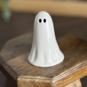 May include: A small, white ceramic ghost figurine with two black dot eyes. The ghost has a simple, flowing design and is sitting on a wooden surface. Ideal for Halloween or spooky-themed decorations.