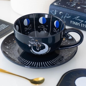 May include: Dark blue ceramic teacup and saucer set with a celestial theme. The cup features moon phase illustrations and the saucer has a crescent moon design with the text "Phases of the Moon". A gold spoon is also visible.