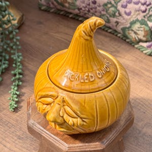 Op de afbeelding: Een gele keramische pot met een deksel in de vorm van een uientop. De pot heeft een gebeeldhouwd gezicht met gesloten ogen en traanvormige details. Het deksel is voorzien van de tekst PICKLED ONIONS in hoofdletters.