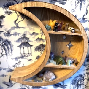 Könnte beinhalten: Ein halbmondförmiges Holzregal mit drei Fächern. Das Regal ist mit kleinen Figuren und Kristallen gefüllt. Der Hintergrund ist eine blau-weiß gemusterte Tapete.