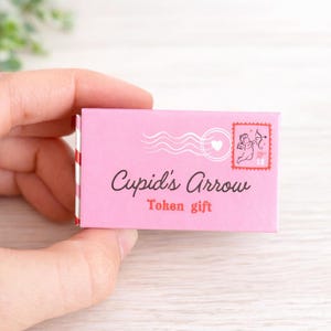 Può includere: Una scatola regalo rosa con le parole "Cupid's Arrow Token gift" stampate sul davanti. La scatola presenta un design a francobollo con un'illustrazione di Cupido e un design a forma di cuore.