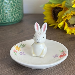 May include: White ceramic ring dish with a small white bunny figurine in the center. The dish is decorated with a floral pattern in shades of pink, purple, yellow, and green.