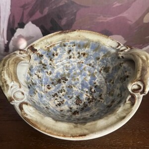 Könnte beinhalten: Eine Keramikschale mit gesprenkeltem blauem Innenraum und hellem braunen Rand. Die Schale hat zwei gebogene Griffe und einen dekorativen Rand. Die Außenseite ist hellbraun.