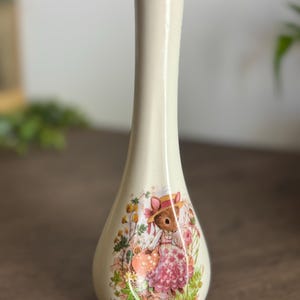 Könnte beinhalten: Cremefarbene Keramikvase mit goldenem Rand und Boden. Die Vase zeigt eine detaillierte Illustration einer Maus mit Hut und Kleid, umgeben von Blumen. Der Text "Friends who share are friends who care" ist auf der Vase aufgedruckt.