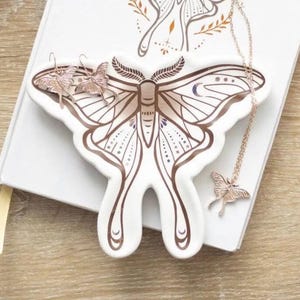 May include: A white ceramic moth-shaped jewellery dish with brown line art. It holds rose gold butterfly earrings and a butterfly pendant necklace. The dish sits on a white book. The earrings and necklace are rose gold.