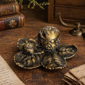 May include: A detailed, antique-style trinket box with a floral design. The box is made of bronze-colored metal and features a spherical lid and a smaller sphere. The base is shaped like a flower with detailed leaf patterns.