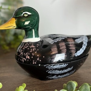May include: A ceramic duck figurine with a glossy finish. The duck has a green head, a yellow beak, and a black body with white and pink accents. The wings are detailed with white brushstrokes. The figurine is likely a decorative item.