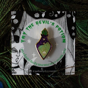 May include: Gold enamel pin featuring a green and purple potion bottle with a skull inside. The pin says "Try the Devil's Potion" and "melancholymemento.com.au".