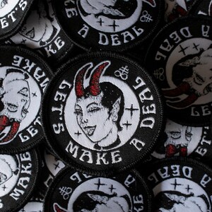 Puede incluir: Primer plano de varios parches bordados en blanco y negro con el texto "Let's Make a Deal" y una imagen de dibujos animados de un diablo con cuernos.