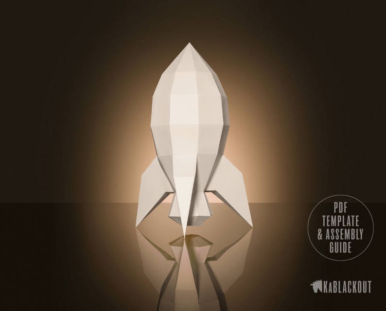 May include: A white paper rocket model with a geometric design. The rocket is standing on a brown surface and is reflected in the surface. The text "PDF TEMPLATE & ASSEMBLY GUIDE" is in a white circle with a black outline. The text "KABBLACKOUT" is in white with a black outline.