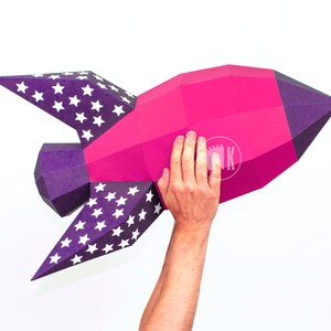May include: A pink and purple paper rocket with white stars on the wings. The rocket is being held up by a person's hand.