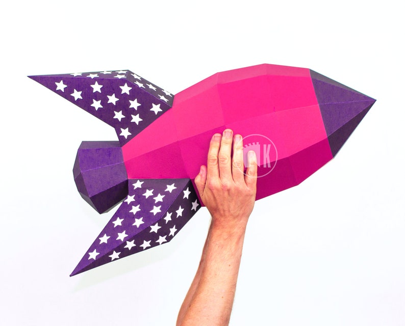 May include: A pink and purple paper rocket with white stars on the wings. The rocket is being held up by a person's hand.