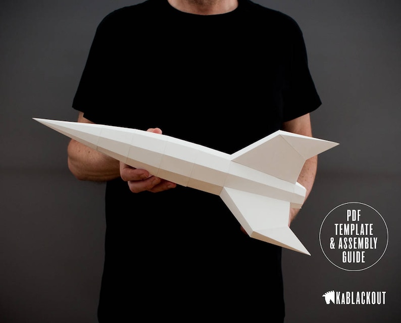 May include: A person holds a white paper model of a rocket ship. The rocket is made of folded paper and has a pointed nose and wings. The text "PDF TEMPLATE & ASSEMBLY GUIDE" is visible in the lower right corner of the image. The text "KABLACKOUT" is visible in the lower right corner of the image.