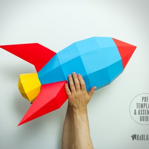 May include: A paper model rocket in blue, red and yellow. The rocket is held up by a hand. The text "PDF TEMPLATE & ASSEMBLY GUIDE" is in a circle with a black background. The text "KABLAKOUT" is below the circle.