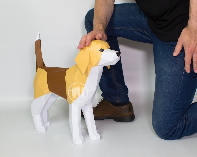 May include: A papercraft model of a beagle dog, standing on a white surface. The dog is white with brown and yellow accents. A person's hand is touching the dog's head.