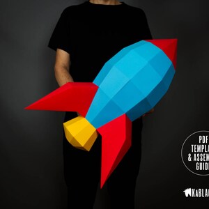 May include: A person is holding a large paper rocket model. The rocket is made of blue, red and yellow paper. The rocket is a 3D model and has a pointed nose and fins. The text "PDF TEMPLATE & ASSEMBLY GUIDE" is visible in the bottom right corner of the image. The text "KABBLACKOUT" is visible in the bottom right corner of the image.