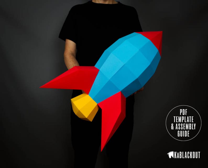 May include: A person is holding a large paper rocket model. The rocket is made of blue, red and yellow paper. The rocket is a 3D model and has a pointed nose and fins. The text "PDF TEMPLATE & ASSEMBLY GUIDE" is visible in the bottom right corner of the image. The text "KABBLACKOUT" is visible in the bottom right corner of the image.