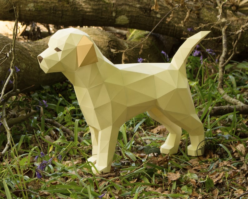 May include: A papercraft model of a yellow Labrador Retriever dog standing in a forest setting. The dog is made of folded paper and has a realistic appearance.