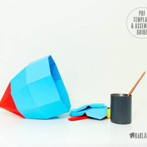 May include: A blue and red paper rocket model with a grey paint can and a paintbrush. The paper model is partially assembled and the paint can is sitting on a white surface. The text "PDF TEMPLATE & ASSEMBLY GUIDE" is visible in the top right corner of the image.
