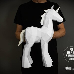 May include: A person holds a white papercraft unicorn model. The model is made of folded paper and has a detailed design. The unicorn is standing on its four legs and has a horn on its head. The person is wearing a black shirt.