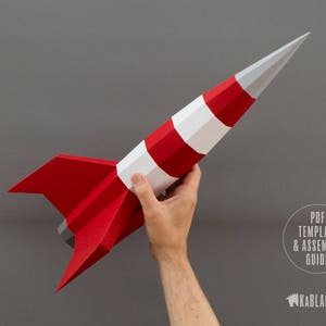 May include: A red and white paper rocket model with a pointed nose and fins. The rocket is held in a person's hand against a gray background. The text "PDF TEMPLATE & ASSEMBLY GUIDE" is visible in the lower right corner of the image.