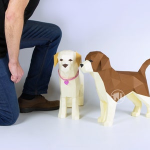 May include: Two papercraft dogs, one brown and white and one white with a pink collar, stand on a white surface. A person's legs and feet are visible in the background.