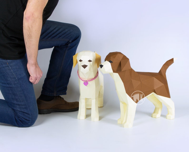May include: Two papercraft dogs, one brown and white and one white with a pink collar, stand on a white surface. A person's legs and feet are visible in the background.