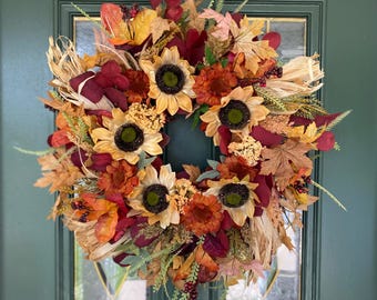 May include: A fall-themed wreath with sunflowers, burgundy leaves, and dried grasses. The wreath is decorated with small red berries and has a rustic, natural look.