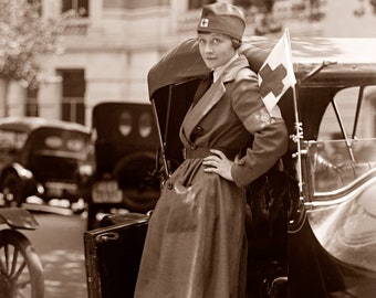 May include: A woman in a brown coat and a red cap with a white cross stands next to a vintage car. She is holding a red cross flag.