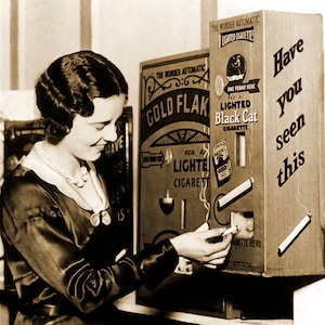 May include: Vintage sepia-toned image of a woman using a cigarette vending machine. The machine has text that reads "Black Cat Cigarettes" and "Have you seen this." The woman is wearing a dark dress and a pearl necklace.